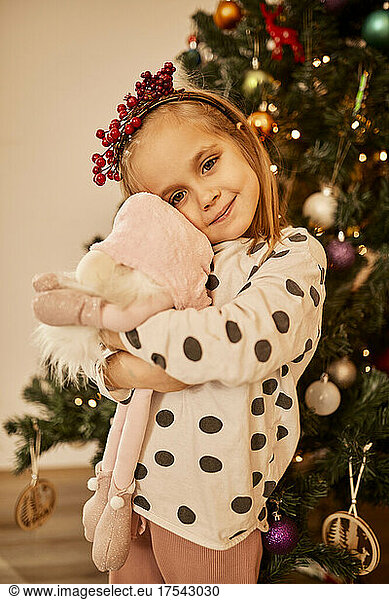 Cute girl embracing stuffed toy standing in front of christmas tree at home