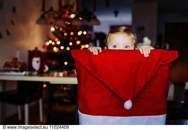 Cute girl hiding behind chair covered with Santa hat at home