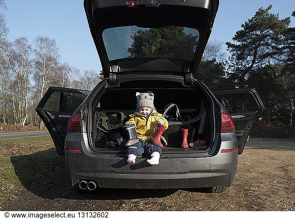 Cute girl holding rubber boots while sitting in car trunk