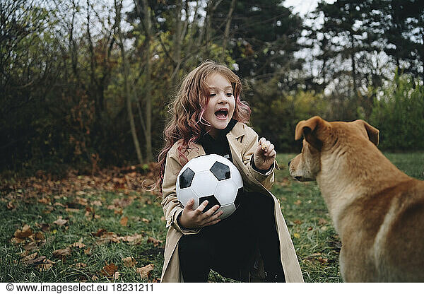 Cute girl holding soccer ball playing with dog at park