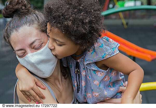 Cute girl kissing her mom at playground outside in a new normal life.