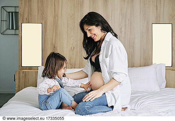 Cute girl touching pregnant mothers belly while sitting on bed at home