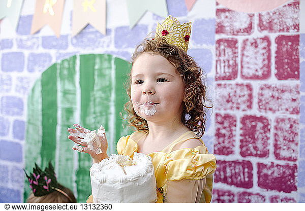 Cute girl with cake standing against castle painting during princess party