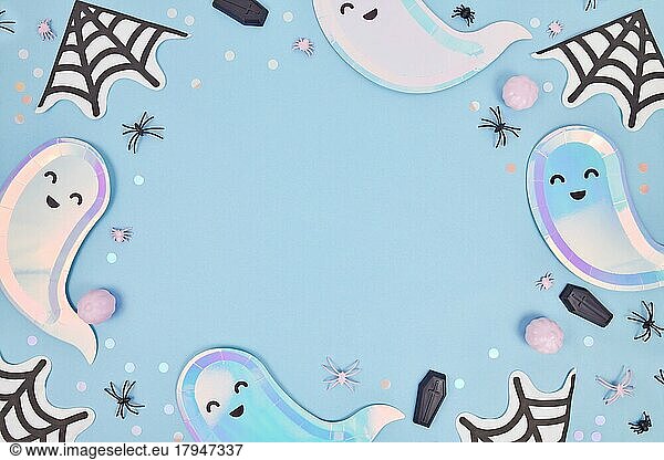 Cute pastel colored Halloween party frame with ghost shaped plates  spider web napkins and confetti on blue background