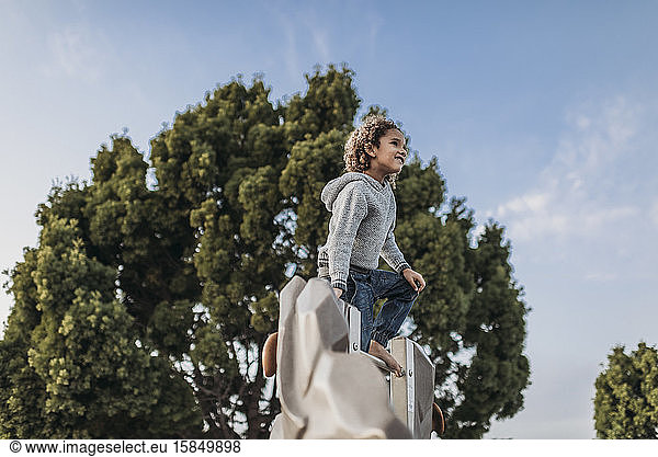 Cute school aged boy standing on top of park structure against trees