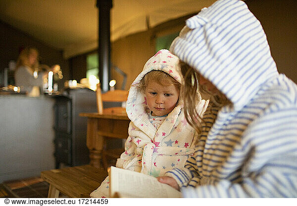 Cute sisters in hooded bathrobes reading book in cabin