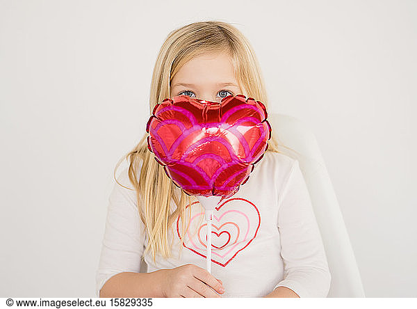 Cute young blond girl holding small red heart balloon under eeditorial