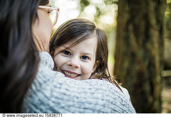 Cute young girl looking over her mothers shoulder in forest.