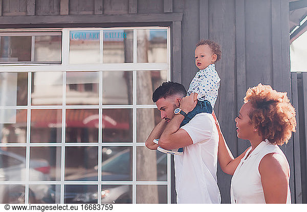 Dad carrying baby on shoulders with mom smiling in front of big window