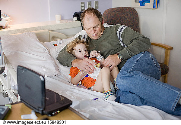 Dad cuddlse with toddler in hospital bed as toddler eats snack.