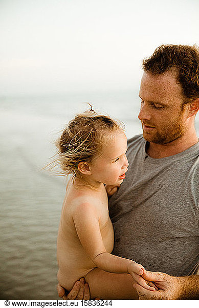 dad in gray shirt holds naked blonde daughter with sunburn on beach