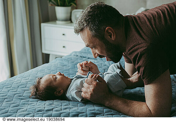 Dad is talking to the baby on the bed holding his hands