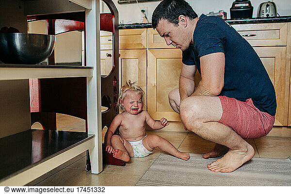 Dad looks down at diapered toddler girl while crying in kitchen