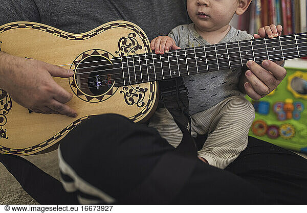 Dad on floor playing child-size guitar while holding 1 yr old on lap.