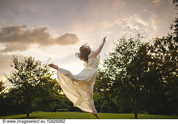 Dancer leaps against cloud-filled sky in park at sunset in gold light