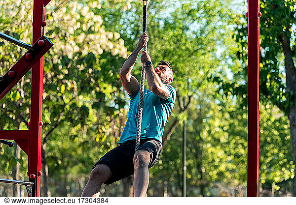 Dark-haired athlete with beard climbing a rope in a park. Outdoor fitness concept.