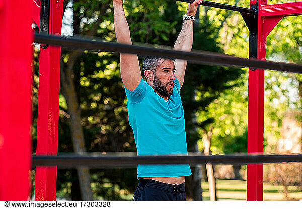 Dark-haired athlete with beard hanging from a calisthenics bar. View between bars. Outdoor crossfit concept.