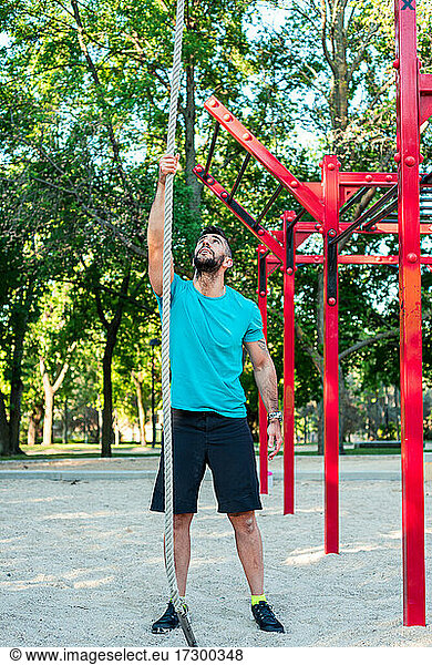 Dark-haired athlete with beard ready to climb a rope. Park background and calisthenics bars.