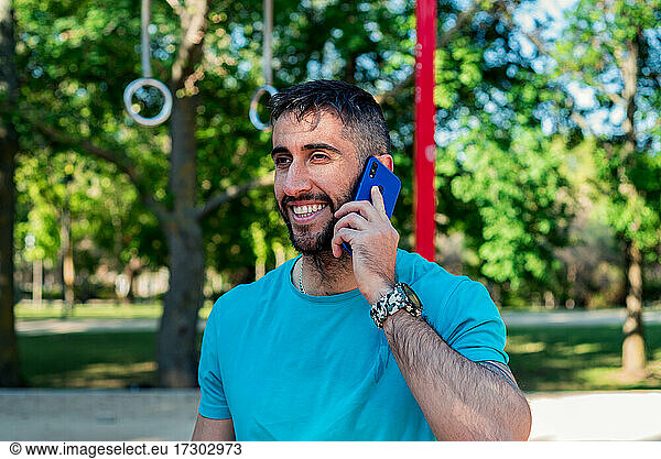 Dark-haired athlete with beard talking on mobile phone after training. Outdoor fitness concept.