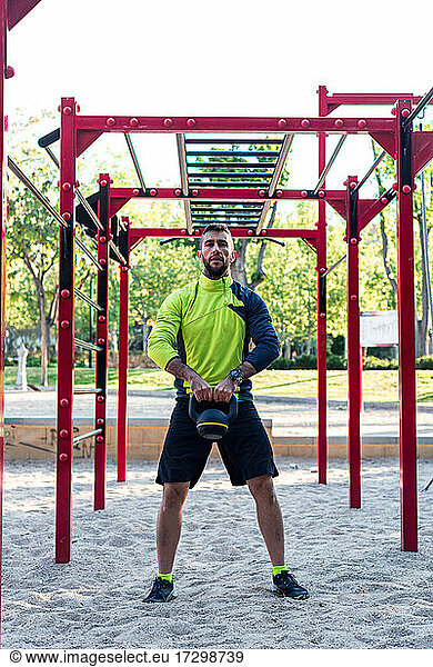 Dark-haired athlete with beard training with kettetbel in a park. Background of calisthenics bars.