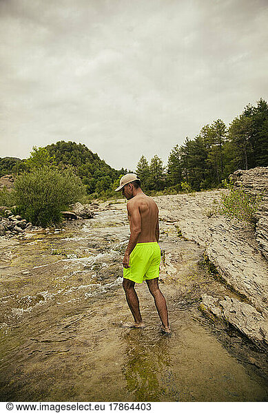 Dark-haired boy in a swimsuit walking in a river between mountains