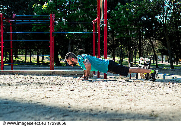 Dark-haired boy with beard doing push-ups in a park. Red fitness bars background.
