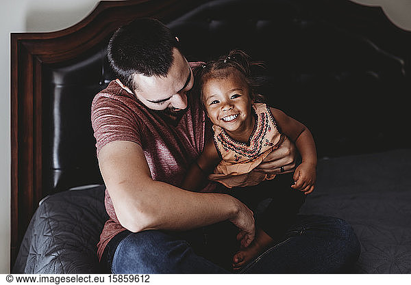 Dark haired dad sitting on bed with smiling multiracial 2 yr old