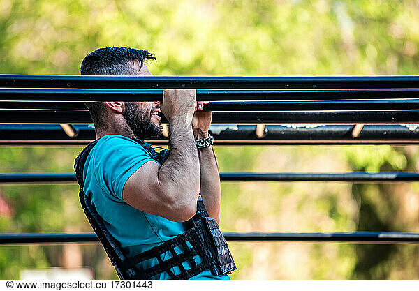 Dark-haired man with beard doing a pull-up on calisthenics bar with weight vest. Outdoor fitness concept.