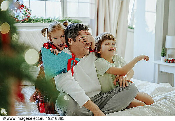 Daughter holding Christmas present and covering father's eyes on bed