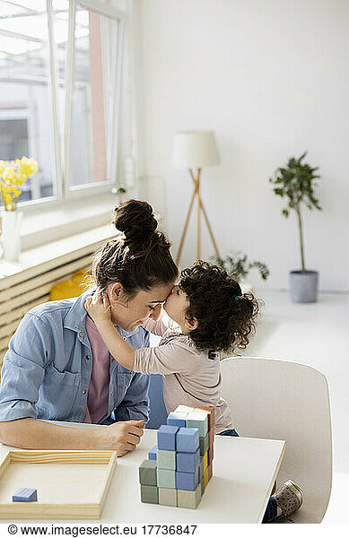 Daughter kissing mother on forehead sitting at table with building bricks