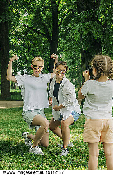 Daughter photographing playful lesbian couple posing with flexing muscles at park