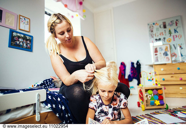 Daughter reading book while mother braiding her hair at home