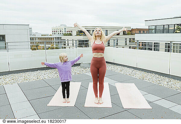 Daughter with arms outstretched looking at mother practicing yoga on rooftop