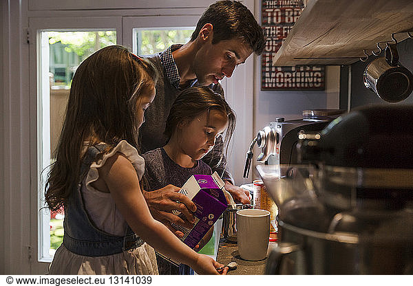 Daughters standing with father pouring milk in mug at kitchen counter