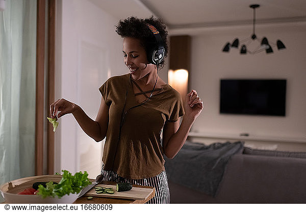 Delighted African American woman in headphones cooking salad