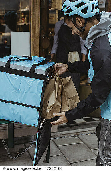 Delivering man putting package order in backpack outside delicatessen shop