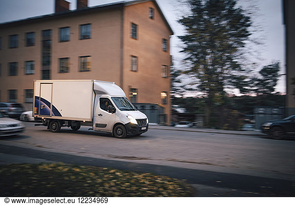 Delivery van moving on road by building in city against sky at sunset