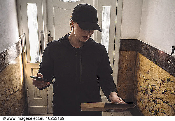 Delivery woman with phone looking at package while standing against door