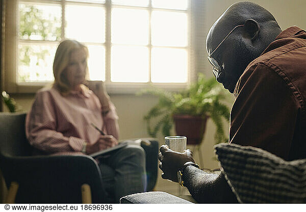 Depressed patient holding glass of water in therapy session with psychologist at office