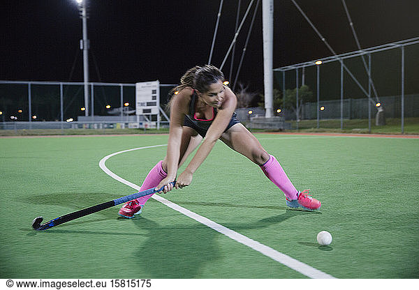 Determined young female field hockey player hitting the ball on field at night