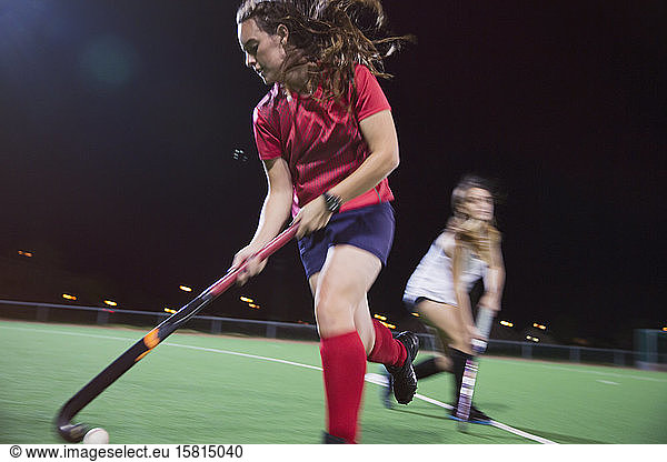 Determined young female field hockey player hitting the ball  playing on field at night