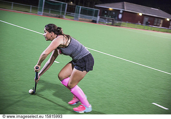 Determined young female field hockey player playing on field at night