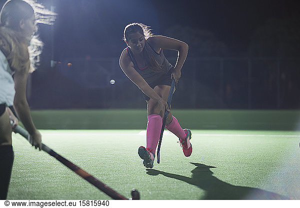 Determined young female field hockey player running with hockey stick  playing on field at night