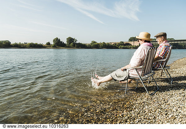 Deutschland  Ludwigshafen  Seniorenpaar sitzt auf Klappstühlen am Flussufer und spritzt mit den Füßen im Wasser.