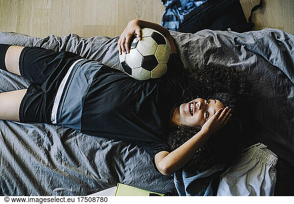 Directly above shot of smiling girl with soccer ball lying down on bed