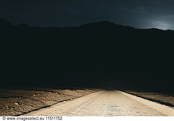 Dirt road in desert illuminated by car headlights  Death Valley National Park  USA  with moonlight in distance.
