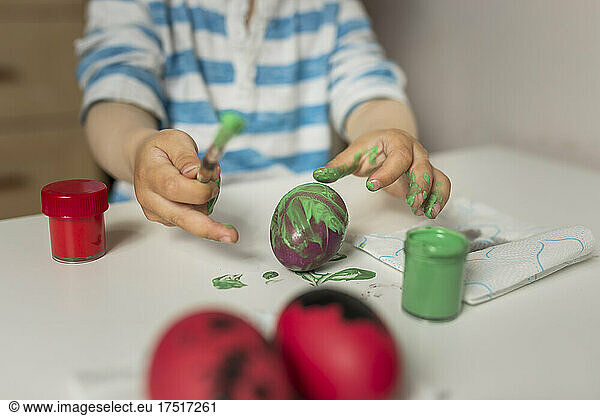 Dirty hands of boy painting easter egg with green paint at home