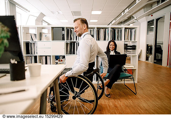 Disabled male professional sitting on wheelchair while smiling businesswoman using laptop at work place