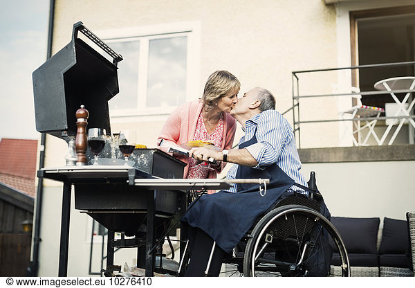 Disabled man in wheelchair kissing woman while barbecuing at yard