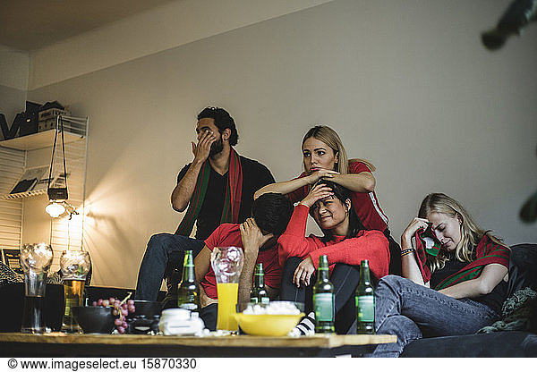 Disappointed fans watching soccer match while sitting on sofa at home
